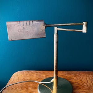 Könnte beinhalten: Eine Schreibtischlampe aus Messing mit rundem Fuß, vertikalem Ständer und verstellbarem horizontalem Arm. Der Lampenkopf besteht aus einem rechteckigen, verwitterten Metallschirm mit einer Reihe vertikaler Schlitze oben und einem weißen Kabel.