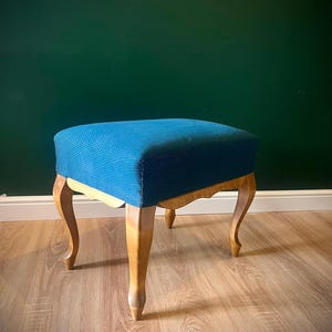 Peut inclure: Un repose-pieds rectangulaire doté d'une assise rembourrée en tissu bleu profond texturé. La base est en bois clair avec quatre pieds incurvés présentant un design de tablier festonné reliant les pieds au cadre de l'assise.