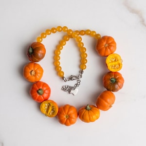 May include: A necklace featuring a strand of translucent yellow beads and several miniature orange pumpkin charms, including two halved pumpkins showing seeds. The necklace has a silver-toned metal lobster clasp and a small heart-shaped charm.