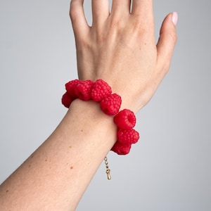 Può includere: Un bracciale caratterizzato da una serie di ciondoli a forma di lampone rosso brillante e realistici, disposti in un motivo circolare. Le singole perle a lampone hanno una superficie ruvida e testurizzata, collegate da una sottile catena in metallo dorato.