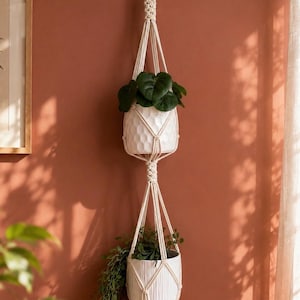 May include: A double-tiered macrame plant hanger made of off-white cotton cord, suspended from a wall hook. It holds two white ceramic pots, one with a textured honeycomb pattern and the other with vertical ridges, each containing a green plant.
