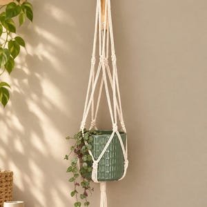 Op de afbeelding: Een crèmekleurige macramé plantenhanger van geknoopt koord, voorzien van een lange kwast aan de onderzijde. De hanger bevat een cilindervormige groene keramische plantenpot met een verticale ribbelstructuur en een hangplant met kleine hartvormige blaadjes.
