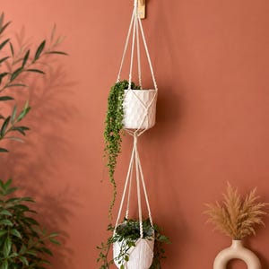 Peut inclure: Une suspension pour plantes en macramé à deux niveaux en corde blanc cassé, avec deux pots en céramique blanche. Le pot supérieur présente un motif hexagonal texturé, tandis que le pot inférieur a une texture côtelée verticale.