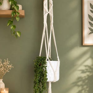 Op de afbeelding: Een crèmekleurige macramé plantenhanger met ingewikkelde knooppatronen en een lange kwast aan de onderkant. De hanger ondersteunt een witte keramische pot met een textuur van zeshoeken, waarin een hangende groene plant met kleine blaadjes staat.
