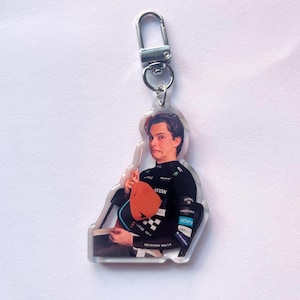 May include: A clear acrylic keyring featuring a printed image of a person with dark hair wearing a black racing suit with various sponsor logos, holding an orange racing cap. The keyring includes a silver-toned metal lobster clasp attachment.