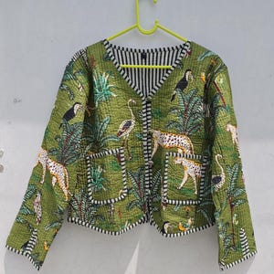 May include: A quilted jacket in olive green featuring a jungle print with leopards, toucans, and flamingos. The garment has a V-neckline, long sleeves, two front patch pockets, and a black and white striped trim along the edges and button placket.