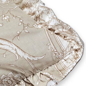 Puede incluir: Un coj&iacute;n cuadrado con una base de tela beige y un patr&oacute;n floral y bot&aacute;nico blanco de estilo toile. El coj&iacute;n est&aacute; rematado con un borde de tela fruncida a lo largo del contorno, que muestra un estampado a juego en blanco y beige.