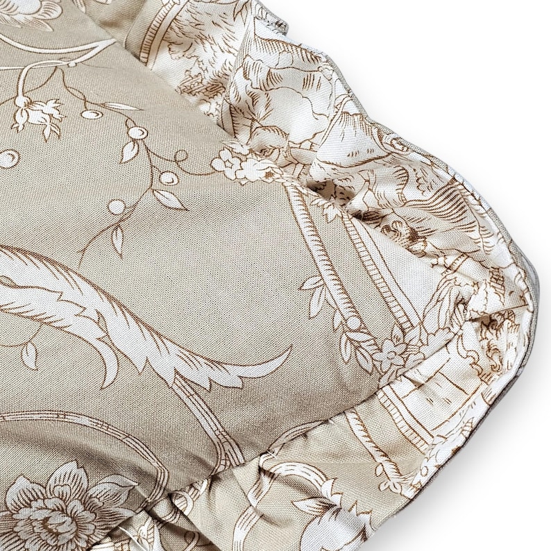 Puede incluir: Un coj&iacute;n cuadrado con una base de tela beige y un patr&oacute;n floral y bot&aacute;nico blanco de estilo toile. El coj&iacute;n est&aacute; rematado con un borde de tela fruncida a lo largo del contorno, que muestra un estampado a juego en blanco y beige.