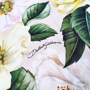 Peut inclure: Un imprim&eacute; sur tissu repr&eacute;sentant des fleurs blanches et jaune p&acirc;le avec des feuilles vertes et le texte Dolce&Gabbana &eacute;crit en noir en cursif au centre. Le motif floral inclut des p&eacute;tales d&eacute;taill&eacute;s et des &eacute;tamines jaunes sur fond clair.