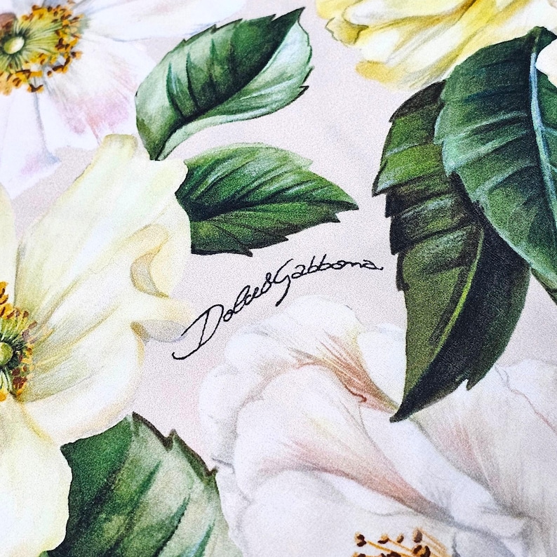 Peut inclure: Un imprim&eacute; sur tissu repr&eacute;sentant des fleurs blanches et jaune p&acirc;le avec des feuilles vertes et le texte Dolce&Gabbana &eacute;crit en noir en cursif au centre. Le motif floral inclut des p&eacute;tales d&eacute;taill&eacute;s et des &eacute;tamines jaunes sur fond clair.