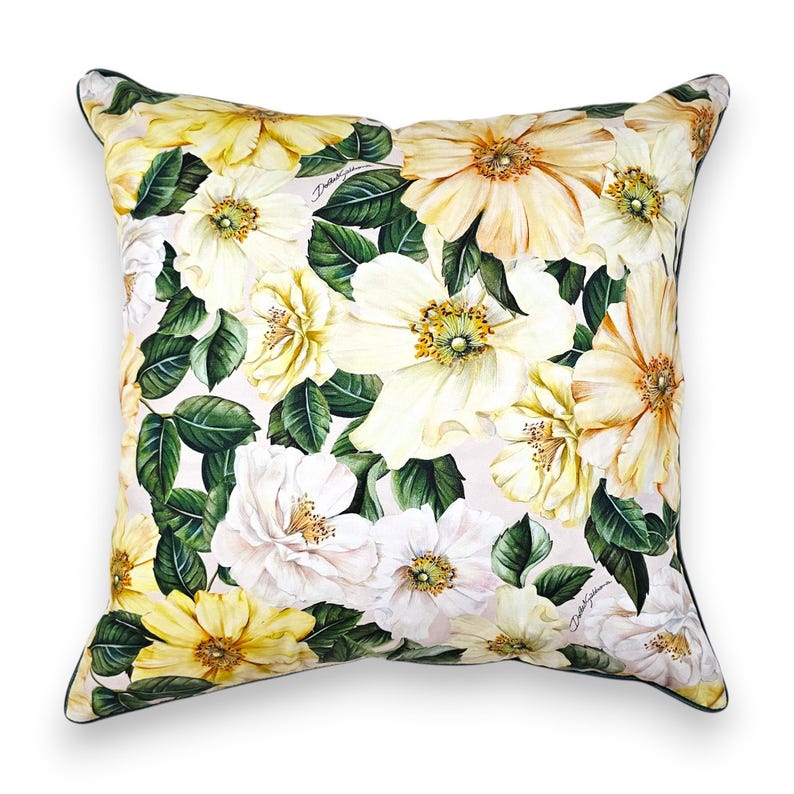 Peut inclure: Un coussin carr&eacute; arborant un imprim&eacute; botanique compos&eacute; de grandes fleurs jaune p&acirc;le et blanches accompagn&eacute;es de feuilles vert fonc&eacute; sur un fond clair. Le tissu pr&eacute;sente le logo Dolce & Gabbana en petites lettres discr&egrave;tes parmi les motifs floraux.
