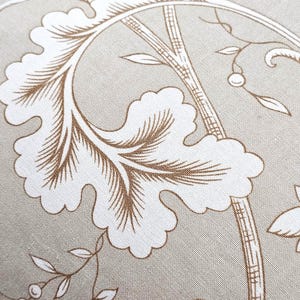 Puede incluir: Una tela de color beige con un estampado bot&aacute;nico blanco que presenta grandes hojas festoneadas y tallos delgados. El patr&oacute;n incluye detalles de l&iacute;neas dentro de las hojas y peque&ntilde;as bayas en ramas delicadas, creando un dise&ntilde;o repetitivo inspirado en la naturaleza.