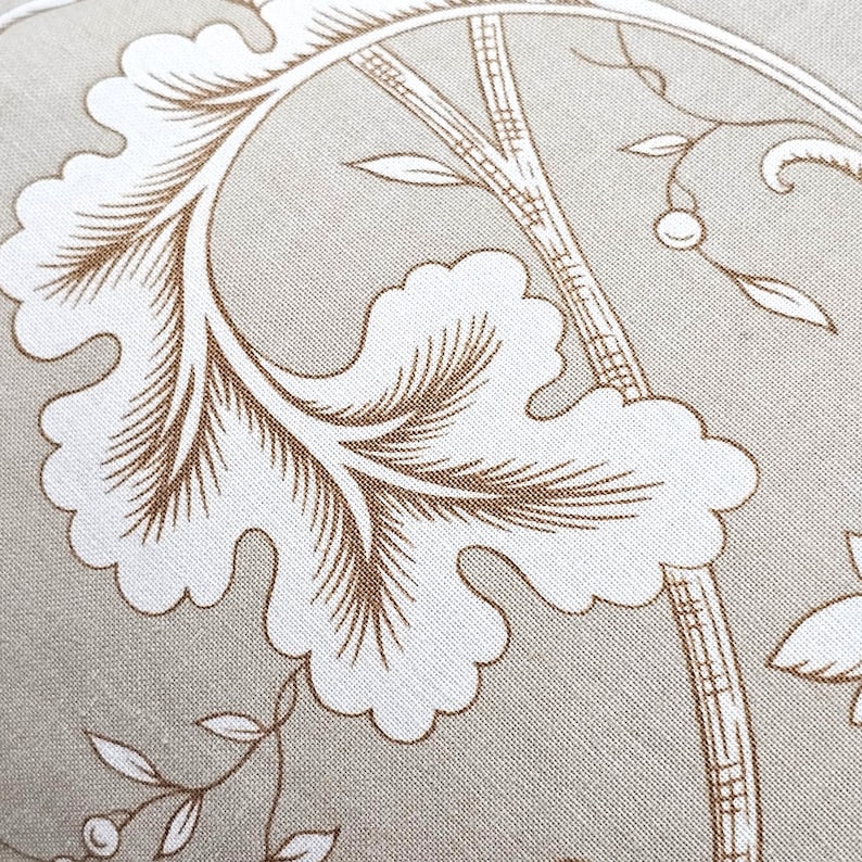 Puede incluir: Una tela de color beige con un estampado bot&aacute;nico blanco que presenta grandes hojas festoneadas y tallos delgados. El patr&oacute;n incluye detalles de l&iacute;neas dentro de las hojas y peque&ntilde;as bayas en ramas delicadas, creando un dise&ntilde;o repetitivo inspirado en la naturaleza.