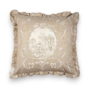 Puede incluir: Un coj&iacute;n cuadrado de tela beige claro con un patr&oacute;n toile de jouy en beige oscuro. El dise&ntilde;o incluye un medall&oacute;n ovalado central que representa figuras en un jard&iacute;n, rodeado de motivos florales y un borde con volantes.