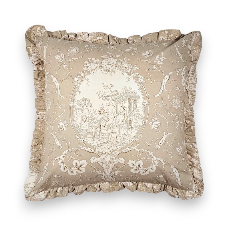 Puede incluir: Un coj&iacute;n cuadrado de tela beige claro con un patr&oacute;n toile de jouy en beige oscuro. El dise&ntilde;o incluye un medall&oacute;n ovalado central que representa figuras en un jard&iacute;n, rodeado de motivos florales y un borde con volantes.