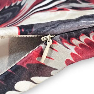Puede incluir: Una vista de cerca de una funda de coj&iacute;n de tela con un patr&oacute;n abstracto vibrante en tonos rojo intenso, negro y blanco roto. La funda incluye un cierre de cremallera oculto con un tirador de pl&aacute;stico beige que muestra el forro interior.