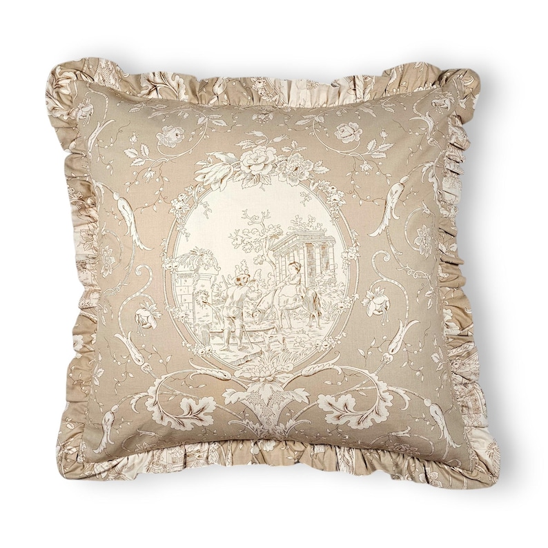 Puede incluir: Un coj&iacute;n cuadrado de tela beige claro con un estampado toile de jouy marr&oacute;n oscuro. El centro presenta un medall&oacute;n ovalado que representa una escena pastoral con figuras y arquitectura, rodeado de motivos florales y un borde con volantes.