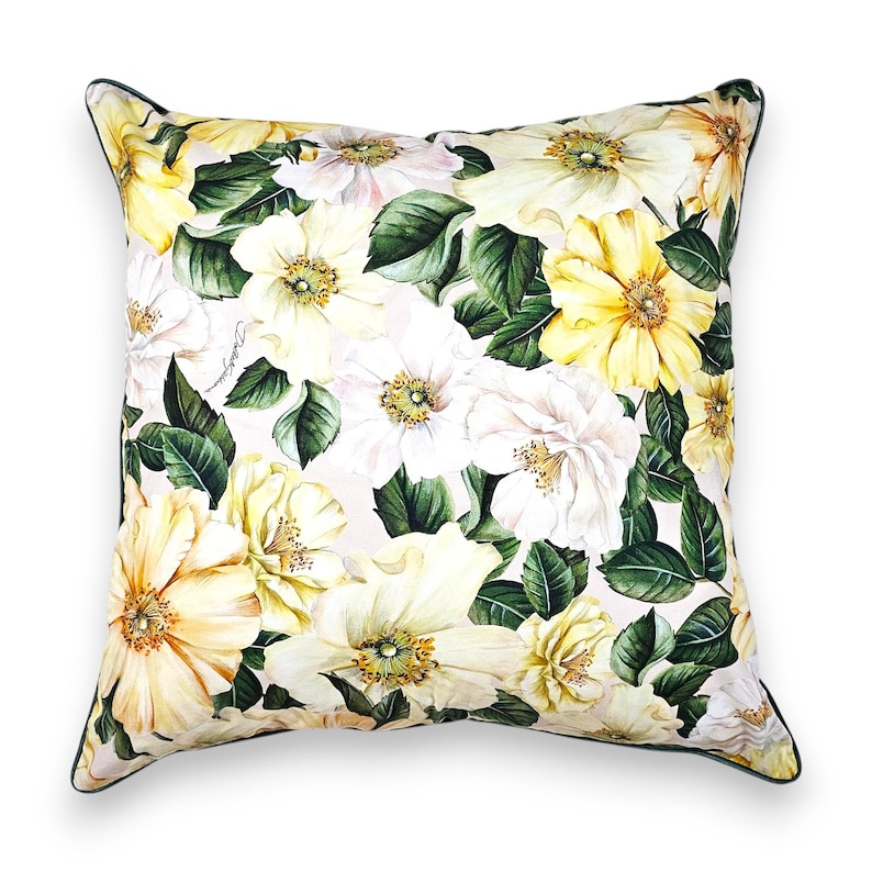 Peut inclure: Un coussin carr&eacute; orn&eacute; d'un imprim&eacute; botanique de fleurs jaunes et blanches avec des feuilles vert fonc&eacute; sur un fond clair. Le coussin pr&eacute;sente un passepoil vert fonc&eacute; et un logo manuscrit signature imprim&eacute; sur le tissu parmi le motif floral.