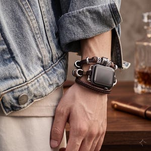May include: A smartwatch with a dark screen featuring a multi-layered brown leather band. The band includes braided leather sections, smooth leather cords, and decorative silver-toned metal rings and filigree beads integrated into the design.