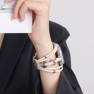 May include: A beige multi-strand watch band featuring braided leather cords and smooth leather strips. The design includes metallic silver-toned beads and circular accents, secured with a metallic clasp attached to a silver-toned smartwatch case.