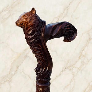 Pode incluir: Um cabo de bengala de madeira castanho-escura esculpido na forma detalhada de uma cabeça e pescoço de urso. A escultura apresenta padrões de pelo texturizados e um acabamento liso e polido, estendendo-se numa pega curva que afunila para uma base circular.