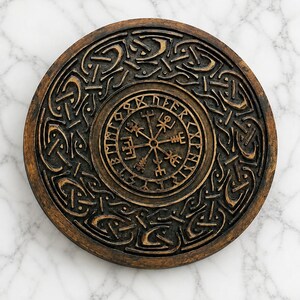 Könnte beinhalten: Eine kreisförmige braune Wandplatte mit einem zentralen Vegvisir-Symbol, das von einem Ring aus Runenzeichen umgeben ist. Der äußere Rand zeigt ein komplexes, erhabenes Knotenmuster mit einem dunklen, verwitterten Finish.