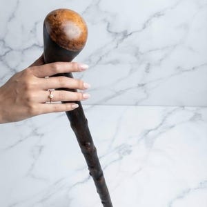Può includere: Un bastone da passeggio in legno marrone scuro con un'impugnatura liscia e arrotondata in radica di legno chiaro. Il fusto del bastone presenta una consistenza naturale, nodosa e irregolare che si restringe verso il basso, mostrando le venature.