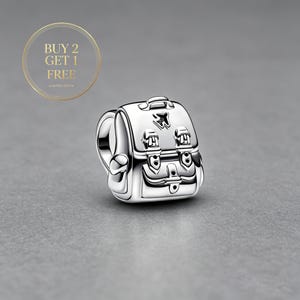 Könnte beinhalten: Ein silberner Charm in Form eines Rucksacks mit zwei Vordertaschen mit Schnallendetails und einem kleinen Flugzeugsymbol auf der oberen Klappe. Der Charm enthält eine kreisförmige Grafik in der oberen linken Ecke mit dem Text BUY 2 GET 1 FREE.