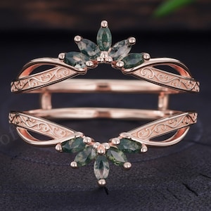 May include: A rose gold ring guard featuring two bands connected by a bridge. The bands are adorned with marquise-cut moss green stones arranged in a fan-like pattern at the centre, complemented by intricate engraved scrollwork along the metal shanks.