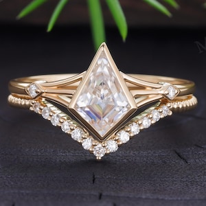 Peut inclure: Une bague en or avec une pierre centrale en forme de cerf-volant sertie clos, accompagnée de petites pierres sur l'anneau. Une alliance en forme de chevron ornée d'une rangée de petites pierres rondes est superposée sous la bague principale.