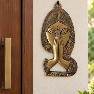 Peut inclure: Une décoration murale couleur laiton représentant un visage stylisé avec les mains jointes en position de prière. La figure présente des cheveux détaillés, des boucles d'oreilles et un bindi. Le mot "Welcome" est gravé au bas de l'objet mural.