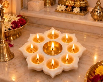 May include: A white marble lotus-shaped oil lamp holder featuring eight outer circular wells and one central brass bowl, each containing a lit cotton wick floating in oil, creating a warm glow on the polished stone surface of the lamp base.