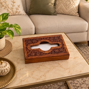 Puede incluir: Una caja rectangular de madera de color marrón oscuro para pañuelos, con intrincados motivos florales tallados a mano en la parte superior. El centro tiene una abertura ovalada alargada con extremos redondeados que muestra un pañuelo blanco.