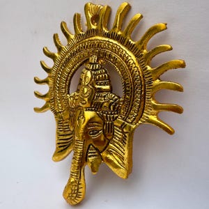 Puede incluir: Una placa de pared de metal dorado que muestra la cabeza de Ganesha con una trompa prominente y orejas grandes. La figura está adornada con una corona ornamentada y enmarcada por un halo radiante en forma de sol con rayos puntiagudos.