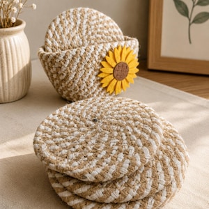 May include: A set of round coasters made from woven tan and white rope, stacked in a matching holder. The holder features a decorative yellow felt sunflower accent on the front. The coasters have a textured, braided appearance with a circular shape.