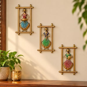 May include: Three rectangular gold-toned metal wall art pieces, each featuring a stylised figure of a person in a colourful outfit. The figures are depicted in different poses, wearing red, blue, and green tops with patterned skirts.
