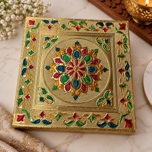 May include: A square gold-coloured box featuring an embossed surface with a central floral mandala pattern. The design is accented with vibrant red, green, and blue enamel-like details, with a textured border framing the central motif.