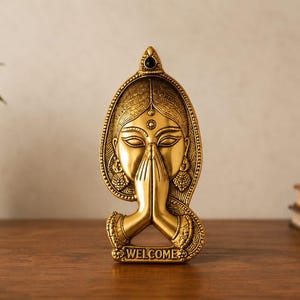 May include: A gold-toned metal wall plaque featuring a sculpted face with closed eyes and hands pressed together in a prayer position. The piece is framed by an ornate archway and includes the word WELCOME embossed at the bottom base.