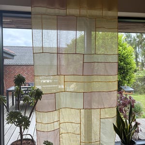 Può includere: Una tenda semitrasparente con un design patchwork composto da pannelli rettangolari in tonalità giallo pallido e rosa tenue. Il tessuto è unito da sottili cuciture visibili che creano un motivo a griglia sull'intera superficie del pannello.