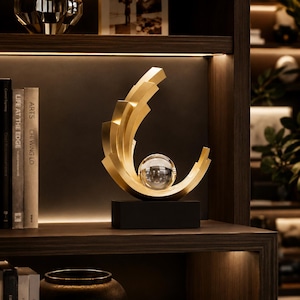 Peut inclure: Une sculpture de couleur dorée composée d'une série de segments métalliques incurvés et étagés disposés en forme de croissant, berçant une sphère en verre transparent au centre, le tout monté sur une base rectangulaire noire solide.