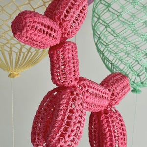 Puede incluir: Una escultura de perro globo tejida a ganchillo en color rosa brillante con una construcción de hilo anudado texturizado. El perro está colocado frente a dos formas de globo esféricas tejidas más grandes en amarillo pálido y verde menta.