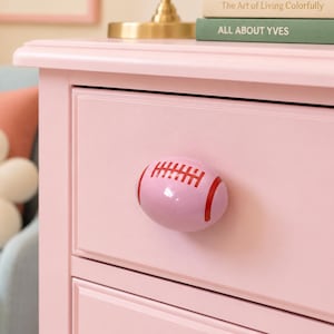 May include: A light pink drawer knob shaped like an American football, featuring a glossy finish with red painted laces and curved end stripes, securely attached to the front of a matching light pink wooden drawer unit with a rectangular frame.