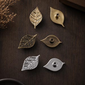 May include: A collection of six leaf-shaped cabinet knobs arranged in pairs. The set includes gold, antique bronze, and silver finishes, each featuring a textured front with visible veins and a smooth back with a central mounting post.