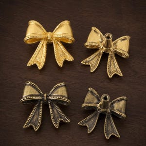 May include: Four metal bow-shaped cabinet knobs in two finishes. Two knobs are bright gold, and two are an antique brass finish. Each knob features a detailed ribbon texture with beaded edges and a central threaded post on the back for mounting.