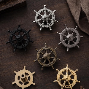 May include: A collection of six ship wheel cabinet knobs in various metallic finishes including silver, black, antique bronze, brushed gold, and polished gold. Each knob features a central hub with spokes radiating to a circular outer rim.