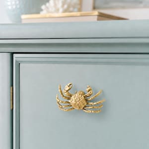 May include: A gold-toned metal cabinet knob shaped like a crab with detailed legs and claws. The crab knob is mounted on a light blue cabinet door with a smooth, matte finish and a visible vertical hinge on the left side of the door frame.