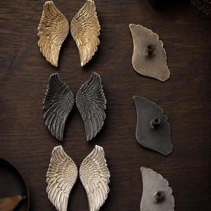 May include: A collection of six wing-shaped cabinet knobs arranged in three pairs. The knobs feature detailed feather textures and come in gold, dark gray, and silver finishes. Each knob includes a central mounting post on the reverse side.