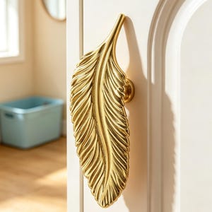 May include: A gold-colored metal door handle shaped like a detailed feather. The handle features a textured surface with a central spine and radiating veins, mounted vertically on a white door panel with a circular base attachment.