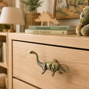 May include: A brass-toned metal drawer knob shaped like a long-necked dinosaur, mounted on a light-colored wooden drawer front. The dinosaur figure features detailed textured skin and a curved tail, serving as a functional handle for the furniture piece.