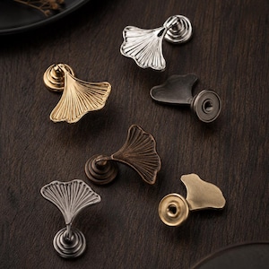 Puede incluir: Una colección de seis pomos de metal para armarios con forma de hojas de ginkgo, con varios acabados metálicos que incluyen oro pulido, plata cepillada, bronce antiguo y negro mate, cada uno con una base circular y un tirador en abanico.